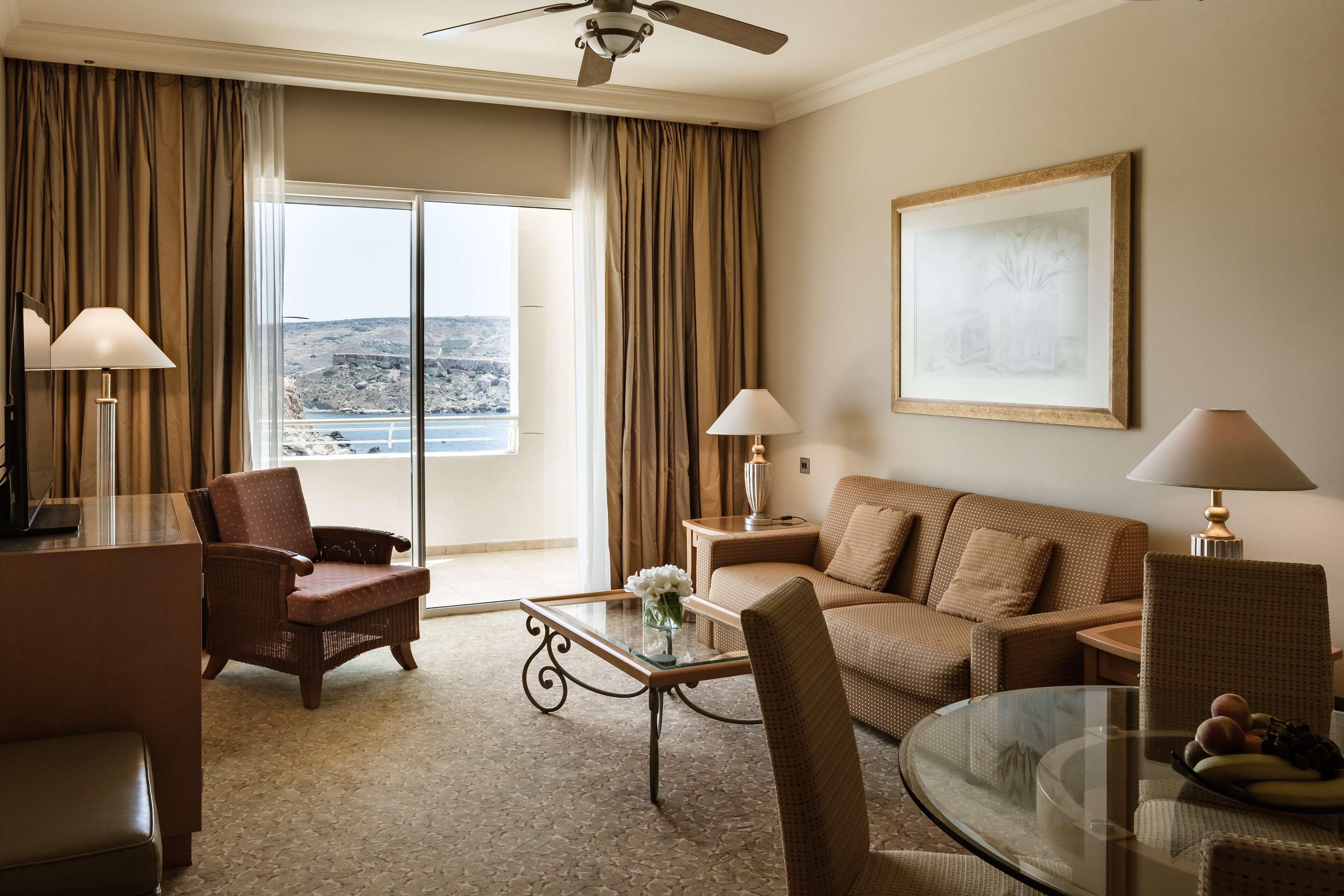Habitación de hotel con balcón, un sofá, una silla, una mesa de centro de vidrio y una lámpara.