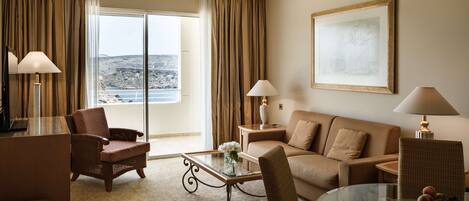 Habitación de hotel con balcón, un sofá, una silla, una mesa de centro de vidrio y una lámpara.