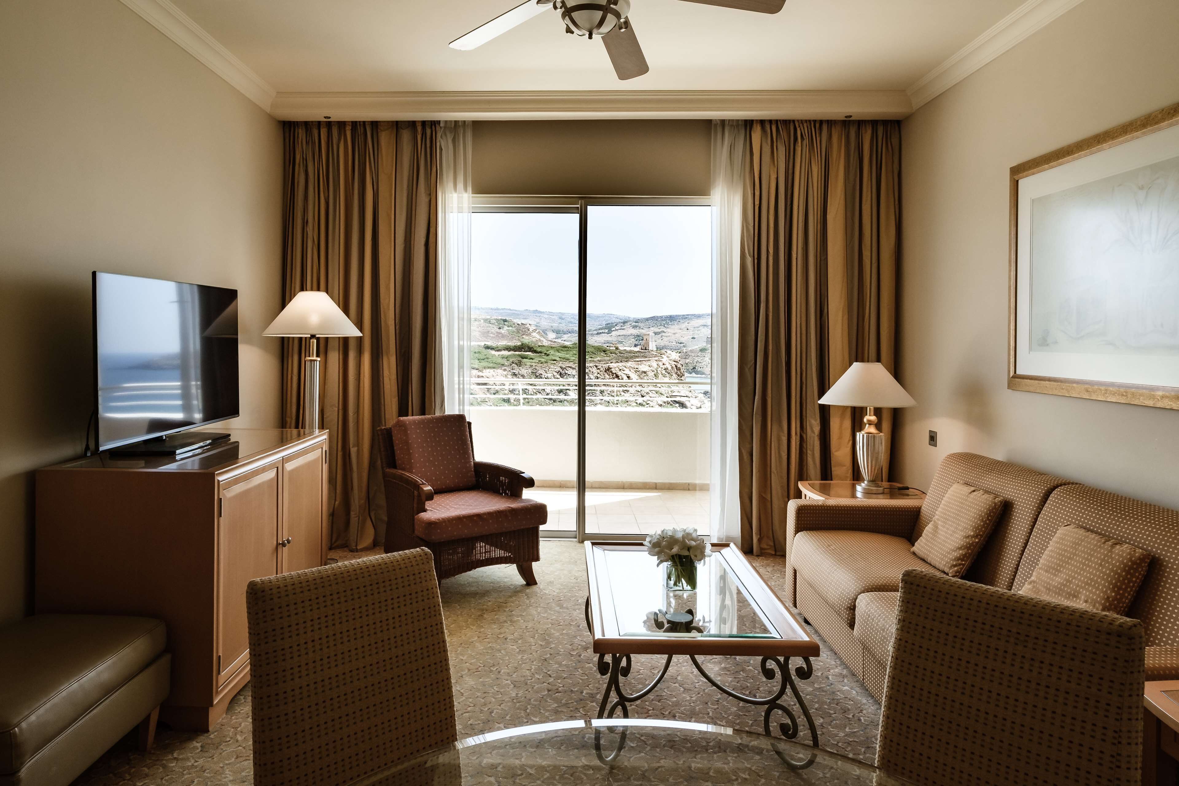 A hotel room with a television, two armchairs, a sofa, a coffee table, and a balcony with curtains.