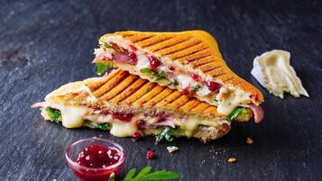 A grilled sandwich cut in half, placed on a dark surface with a small bowl of red condiment and some green leaves nearby.