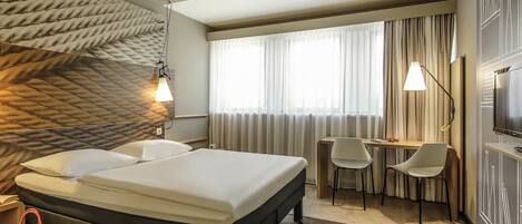 A hotel room with a bed, a desk, two chairs, a TV, and a patterned wall.