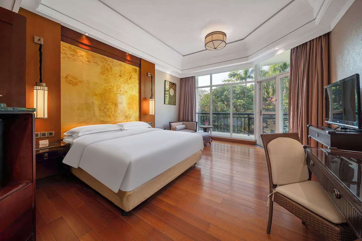 A spacious hotel room with a large bed, a television, a balcony with a view of greenery, and a wicker chair.
