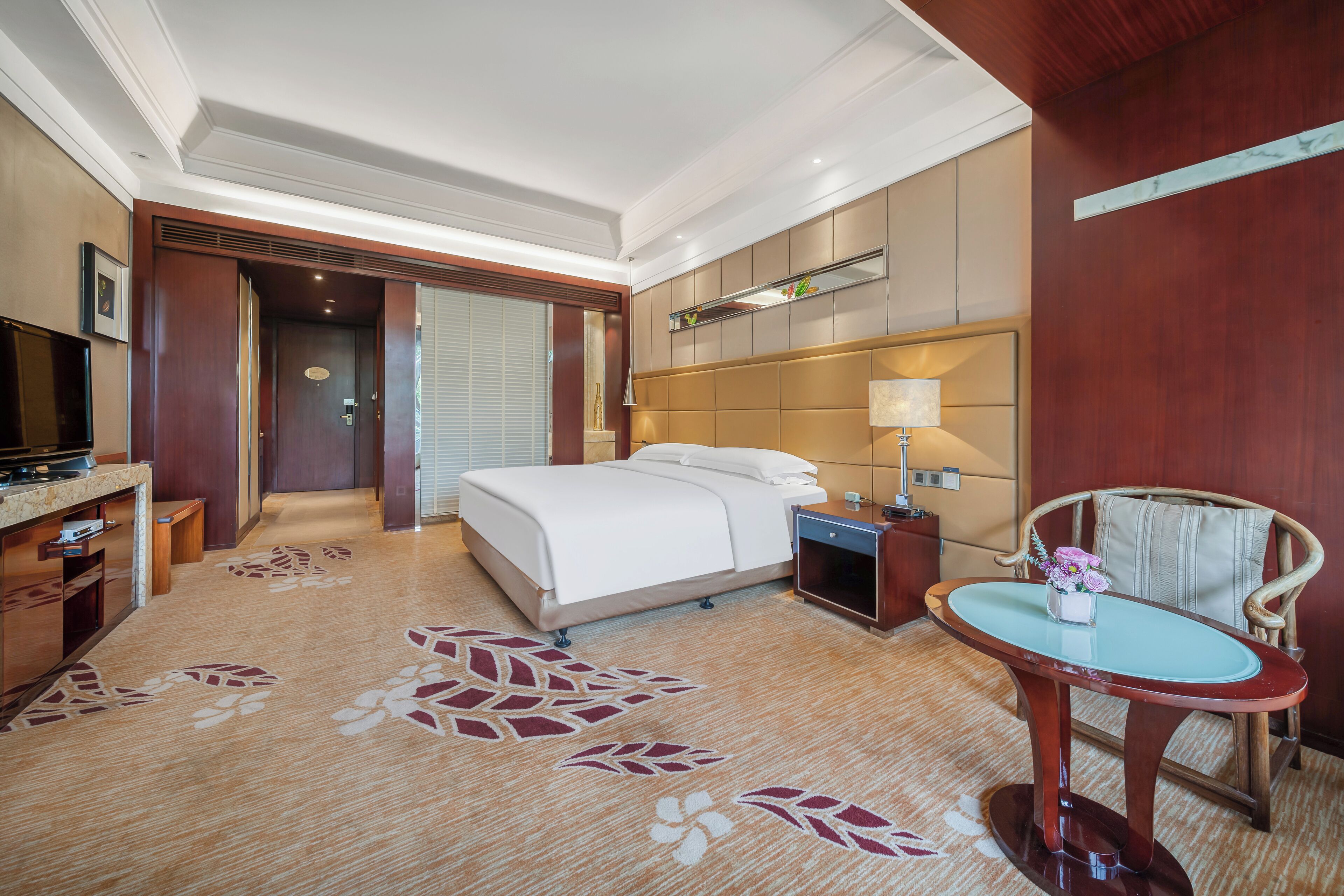 A hotel room with a bed, bedside table, lamp, chair, and a small table with flowers.