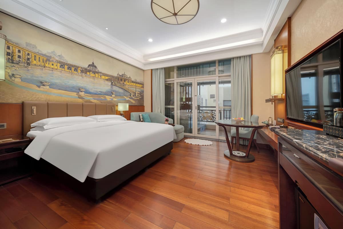 A hotel room with a large bed, a dining table, a TV, and a balcony with a view.