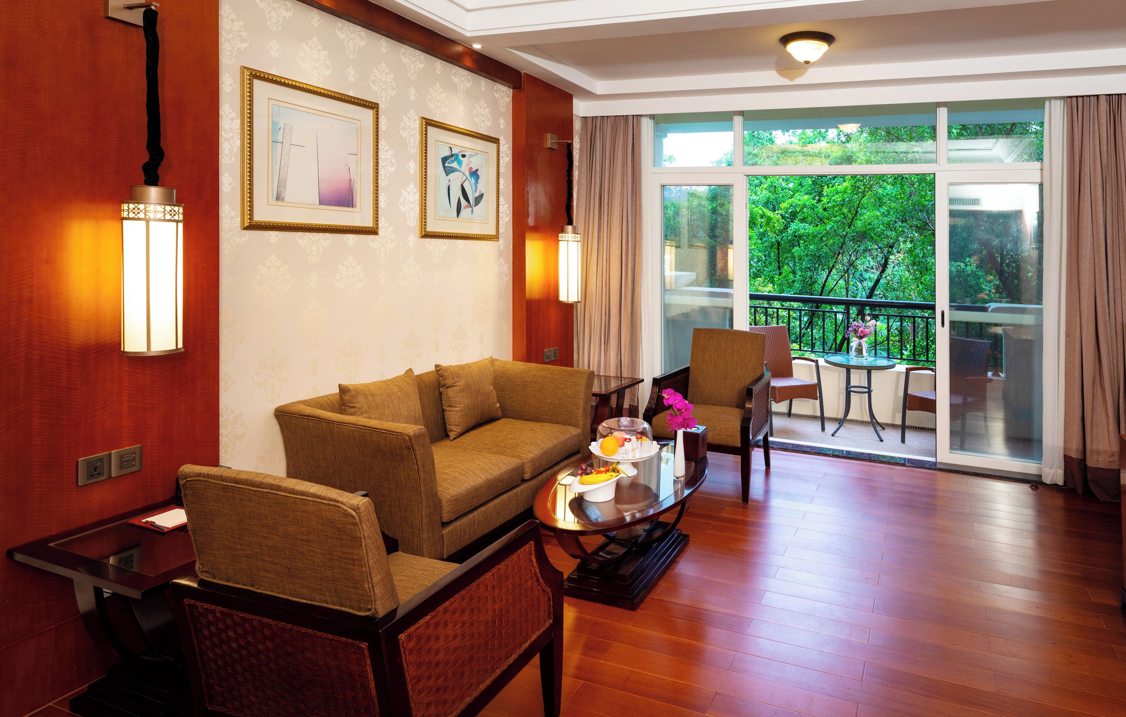 Habitación de hotel con piso de madera, un sofá, una mesa de comedor con un jarrón y flores, y un balcón con vista a árboles.