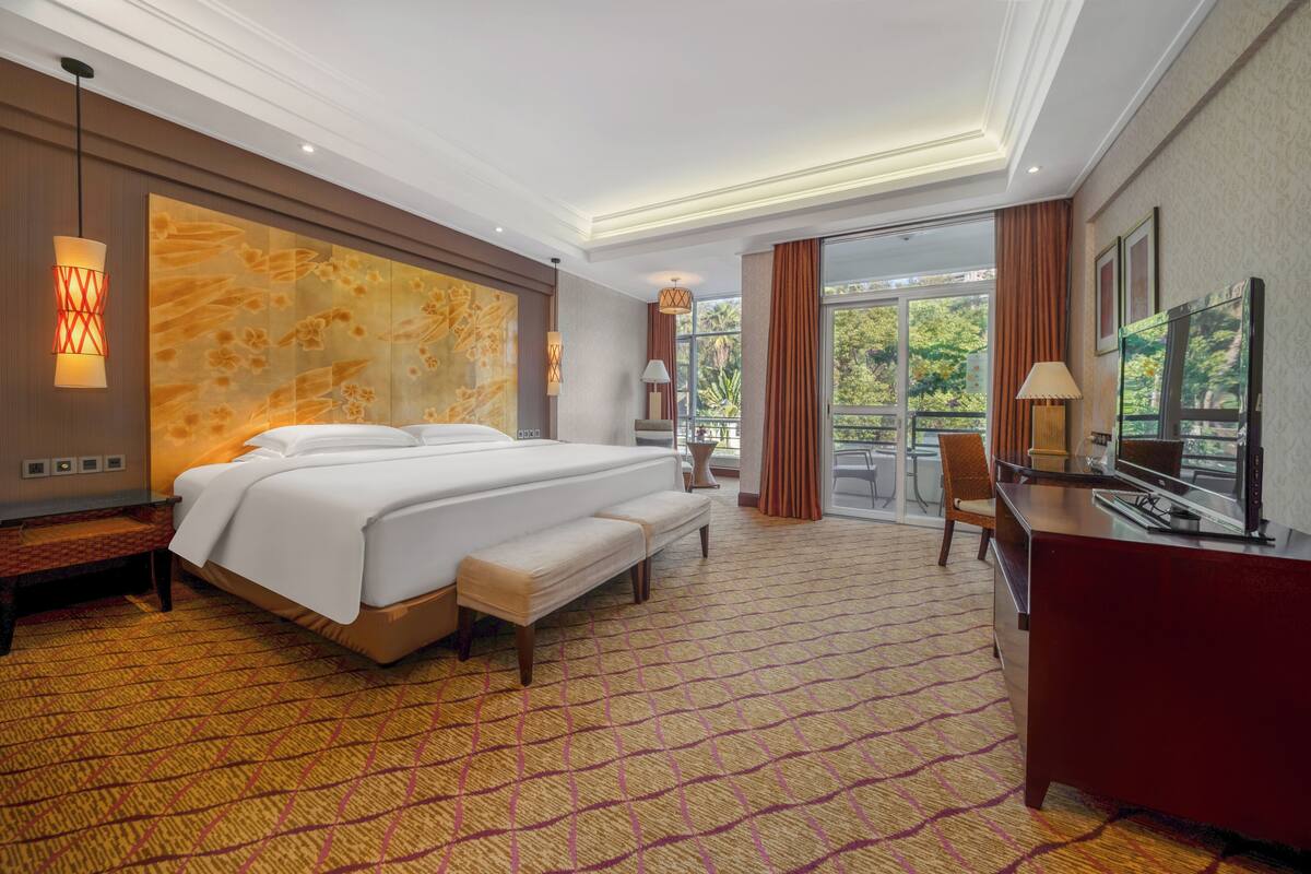 A hotel room with a large bed, a desk, a chair, and a balcony with a view of greenery.