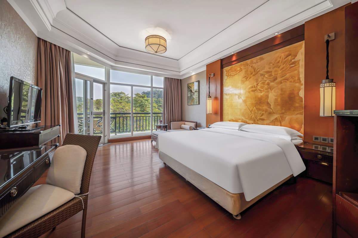 A hotel room with a large bed, a television, a desk, and a balcony with a view of greenery.
