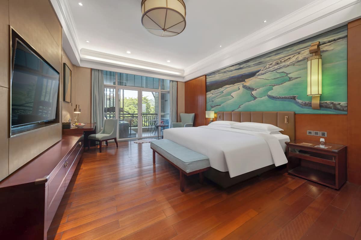 A modern hotel room with a large bed, a flat-screen TV, a seating area with a table and chairs, and a balcony with a view.