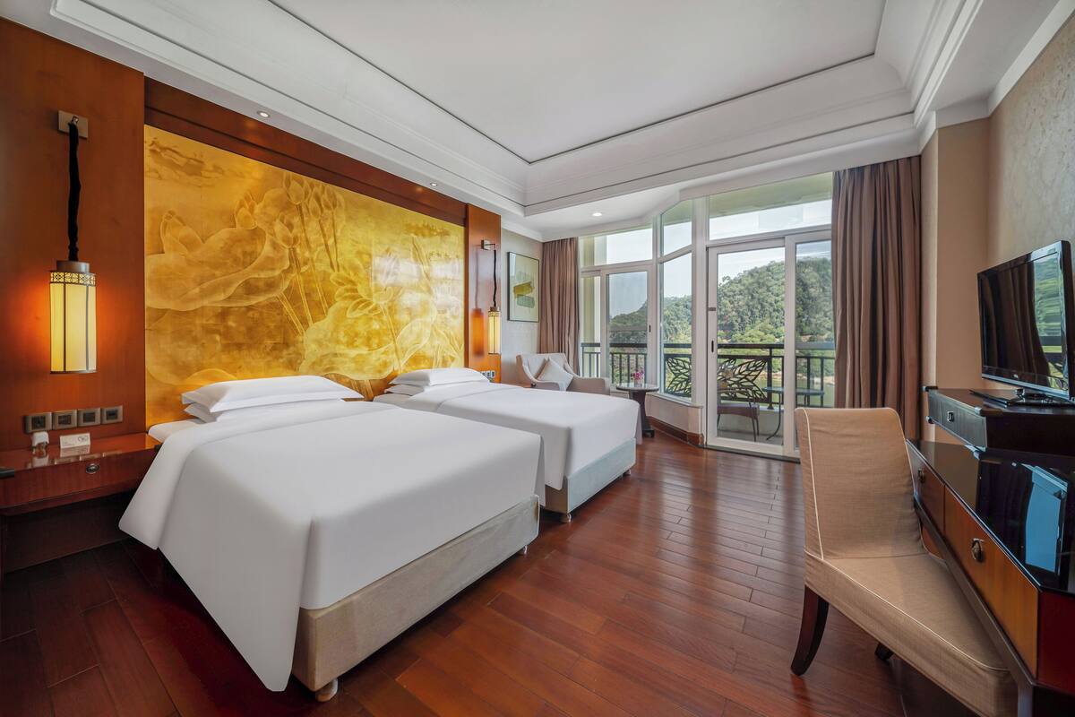 A hotel room with two beds, a TV, a desk, and a balcony with a view.