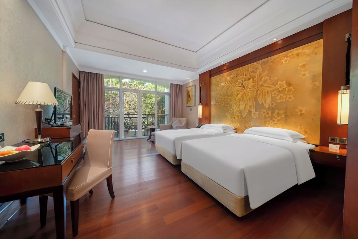 A hotel room with two beds, a desk with a lamp, a chair, and a view of greenery through the window.