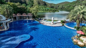 Una gran piscina azul cristalina con una estatua en el centro, rodeada de palmeras y un lago al fondo.