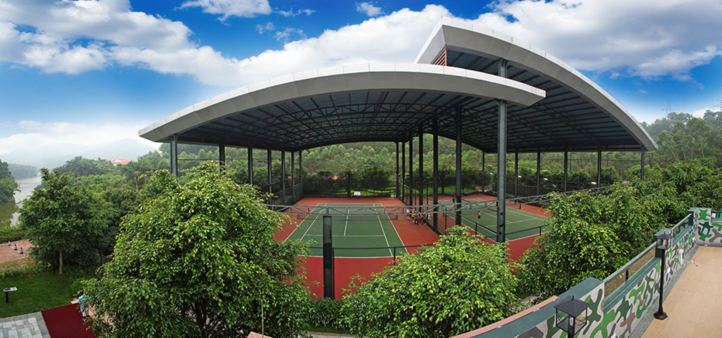 Un campo da tennis con una tribuna coperta, circondato da alberi e un cielo sereno.