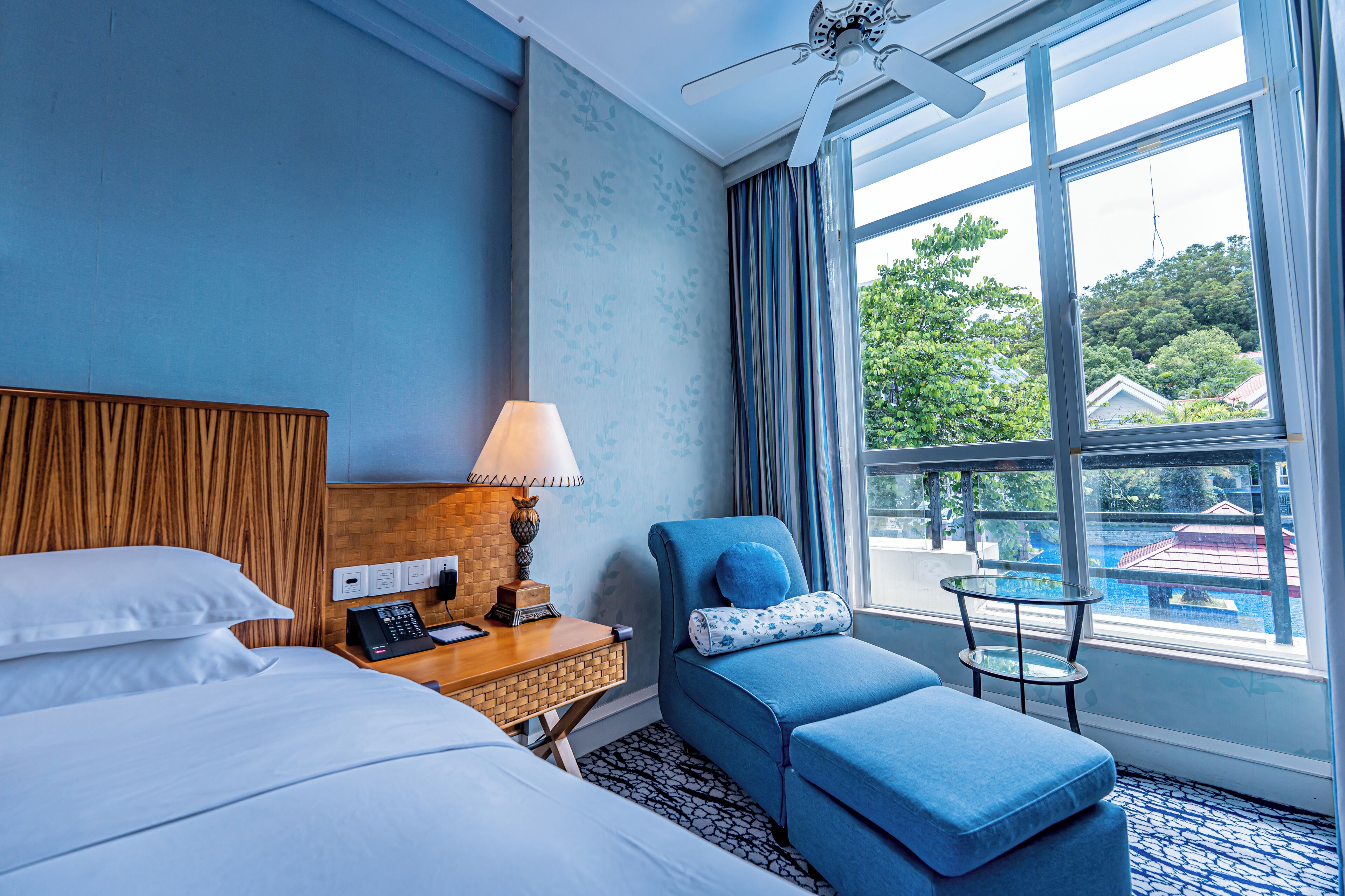 Habitación de hotel con cama, escritorio con lámpara, zona de estar con cojín y vistas a una piscina a través de amplias ventanas.