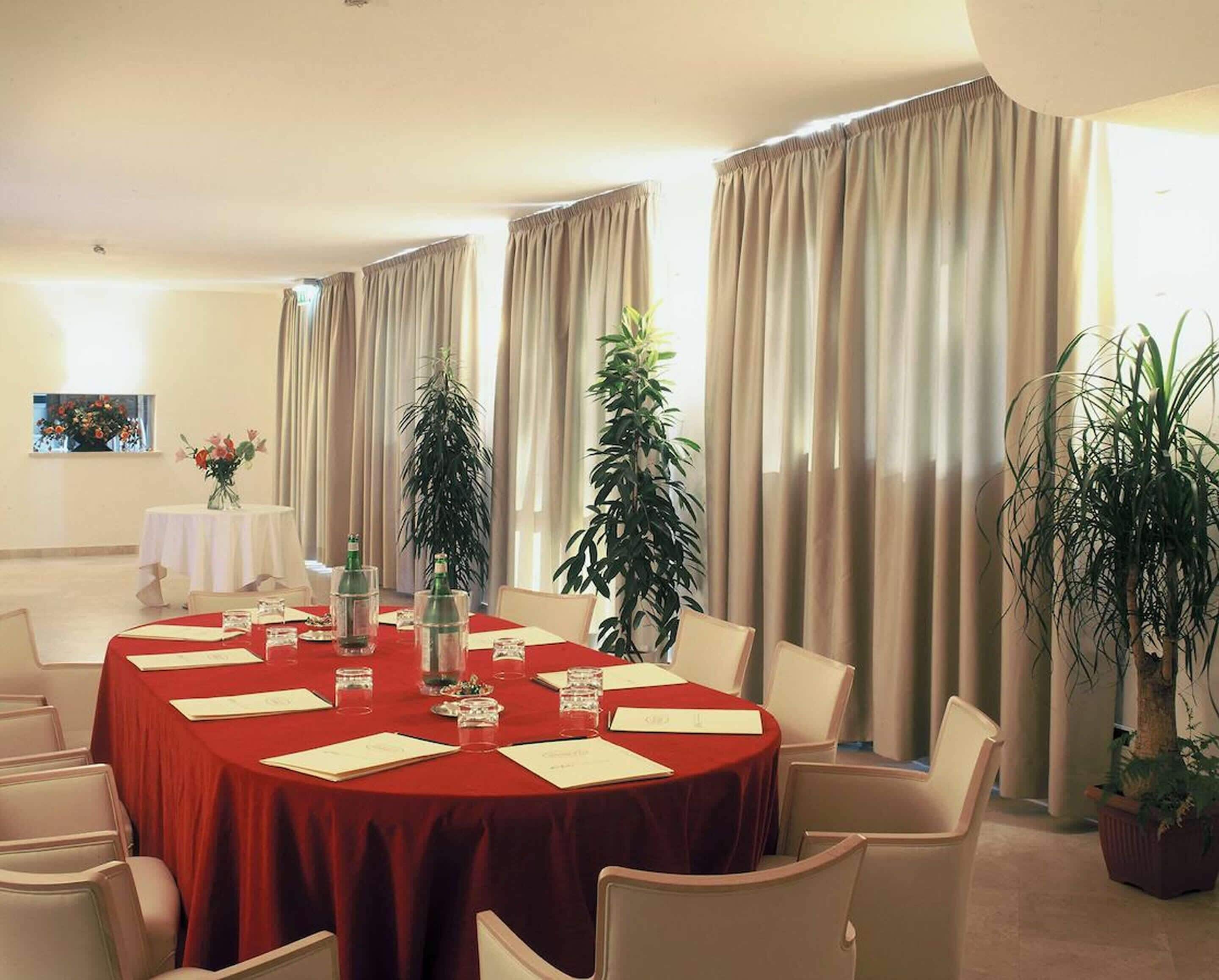 Une salle de conférence avec une table ronde recouverte d’une nappe rouge, entourée de chaises blanches. De part et d’autre de la table, on aperçoit des plantes en pot, et au fond, une fenêtre avec des rideaux.