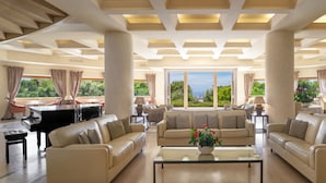 A spacious living room with a grand piano, comfortable sofas, and a large window offering a scenic view. - Tombolo Talasso & Beach Resort (Castagneto Carducci)