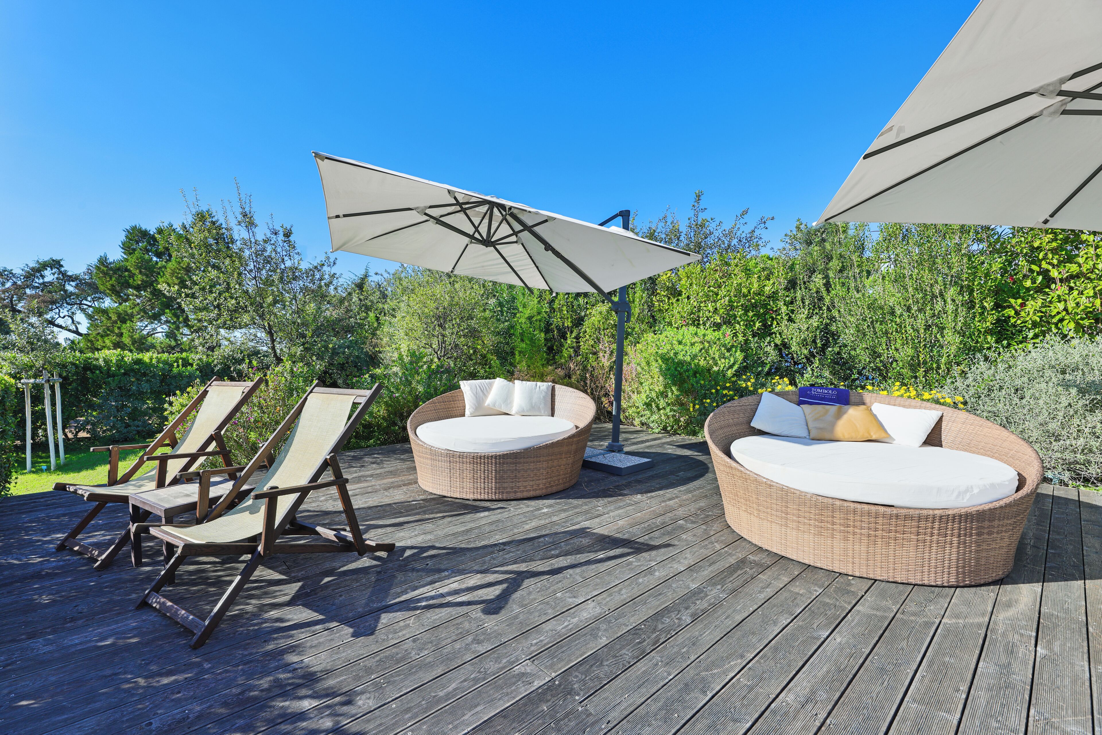 Una terraza de madera con mobiliario de mimbre, incluyendo dos sillas redondas y un sofá, bajo grandes sombrillas blancas.