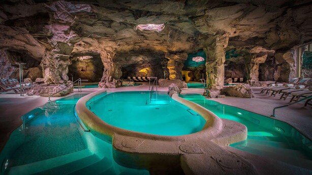 An indoor pool with a natural rock backdrop, surrounded by lounge chairs and a stone pathway. - Tombolo Talasso & Beach Resort (Castagneto Carducci)