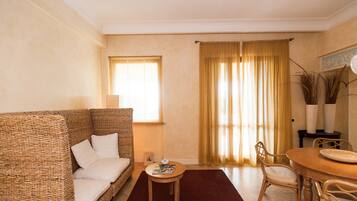 A living room with a wicker sofa, a small round table, a dining table with chairs, and a window with curtains.