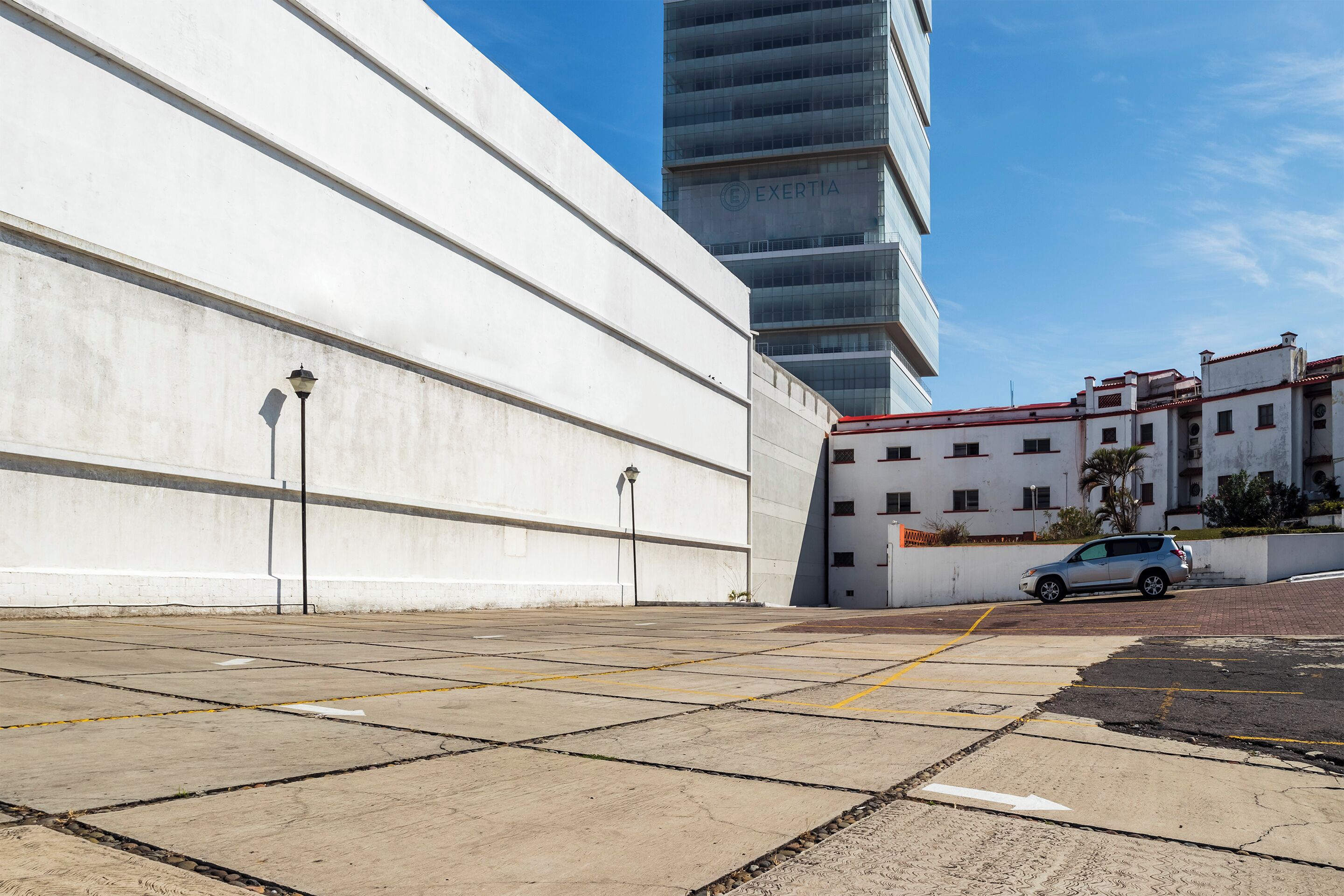A parking area with a white building and a modern high-rise in the background.