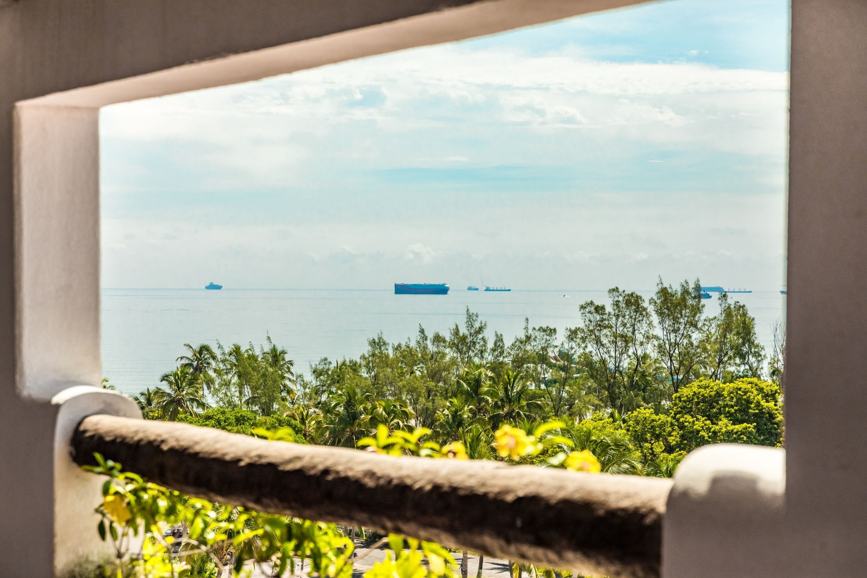 Vista a través de una ventana con barandilla de cuerda, sobre un mar tranquilo con barcos y abundante vegetación.