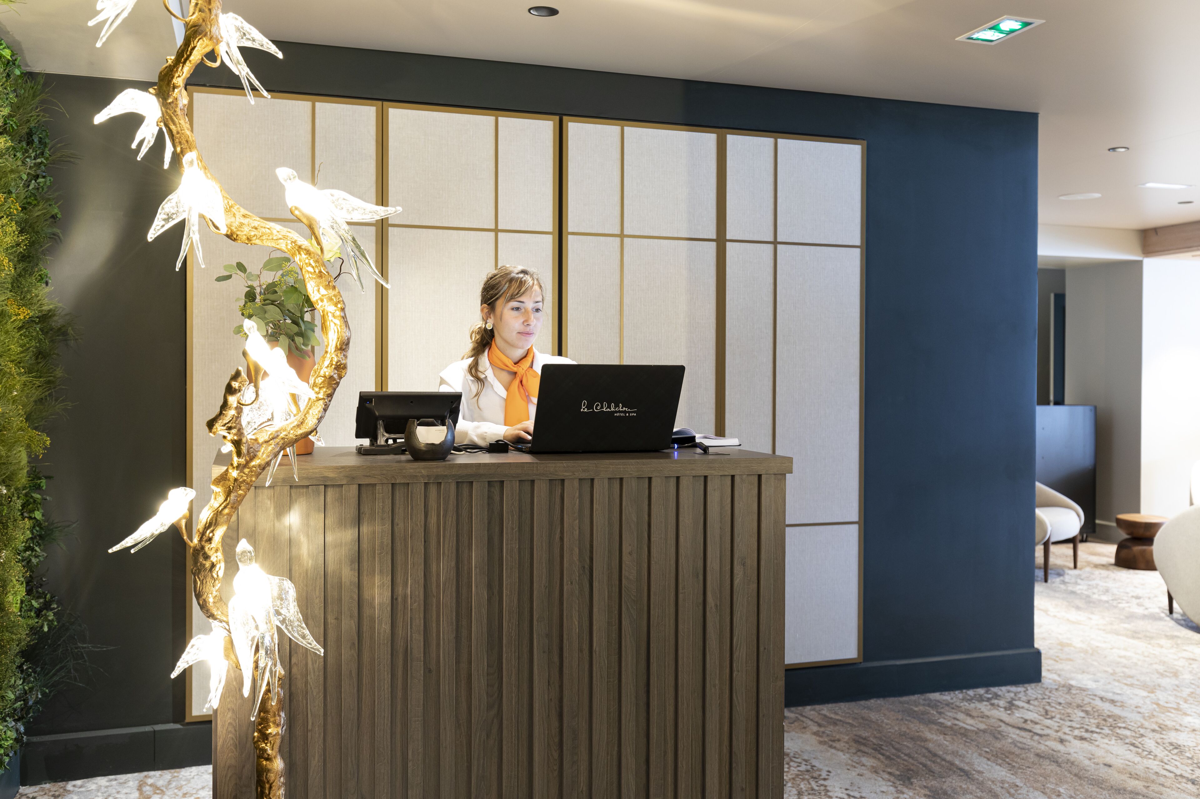 Un comptoir d’accueil avec un ordinateur portable, un arbre décoratif illuminé et un espace salon en arrière-plan.