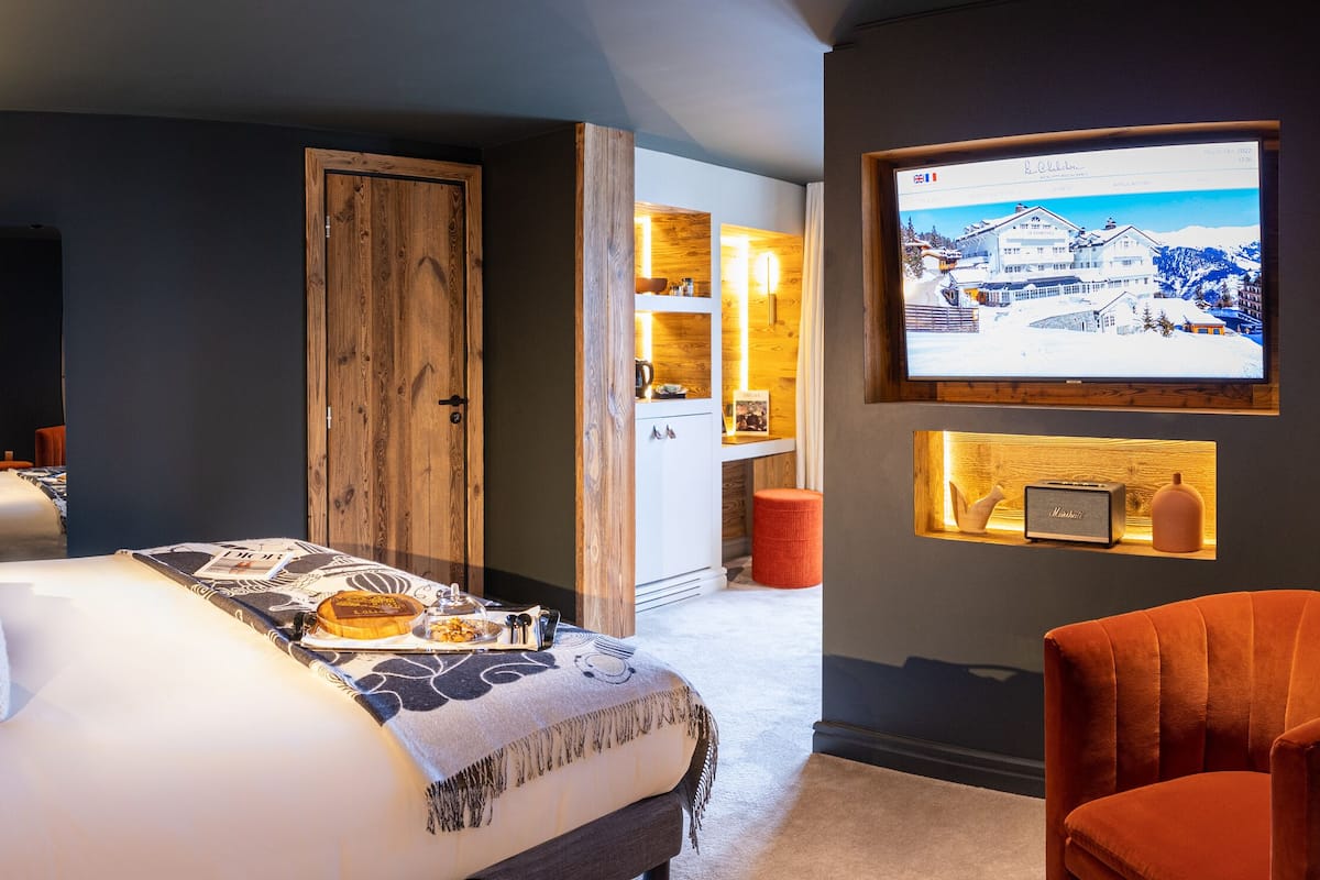 Quarto de hotel moderno com uma cama grande, televisão e uma poltrona laranja aconchegante.