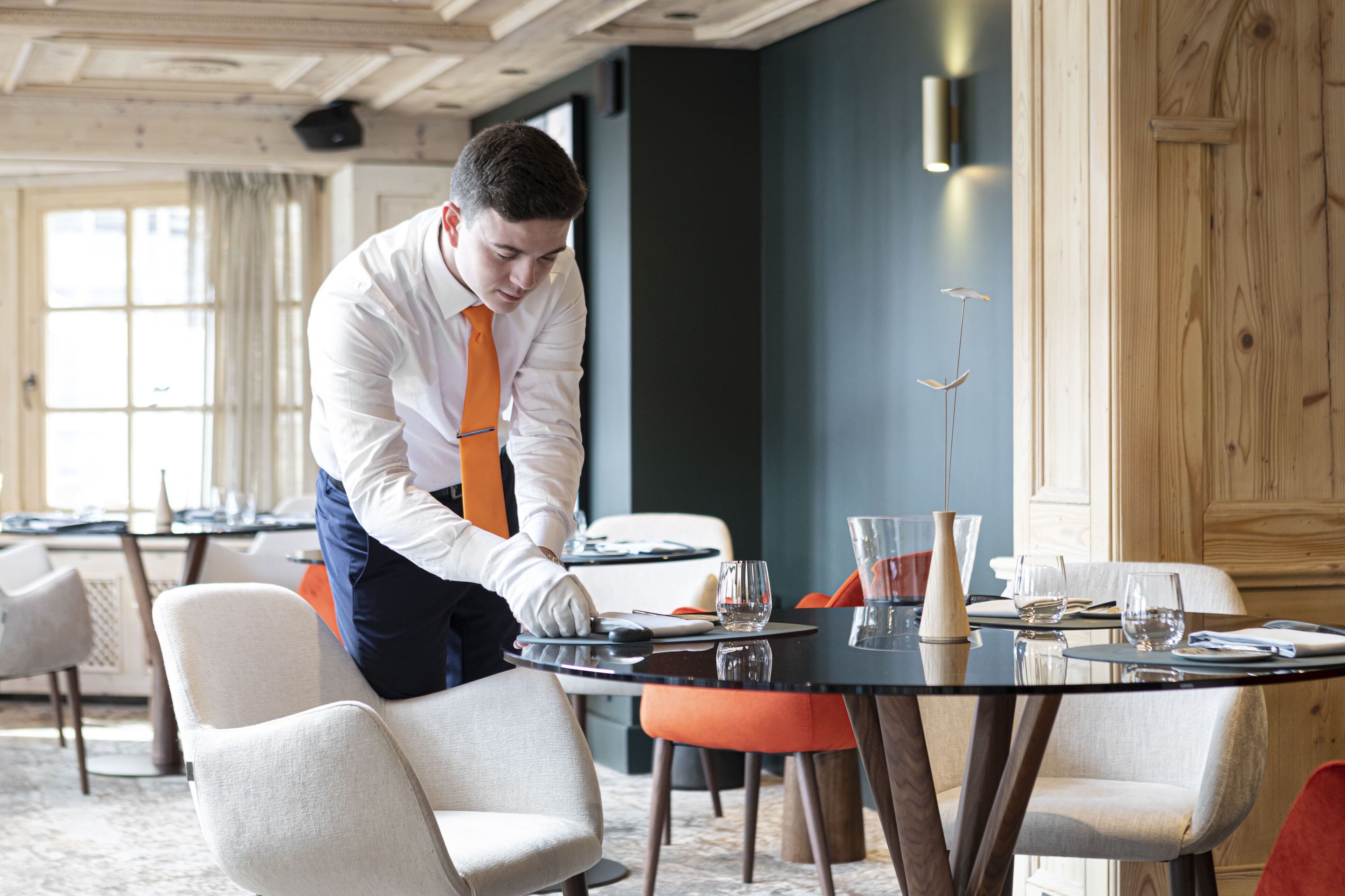 Une salle à manger d’hôtel où un serveur prépare une table. On y trouve plusieurs tables, des chaises et une grande fenêtre qui laisse entrer la lumière naturelle.