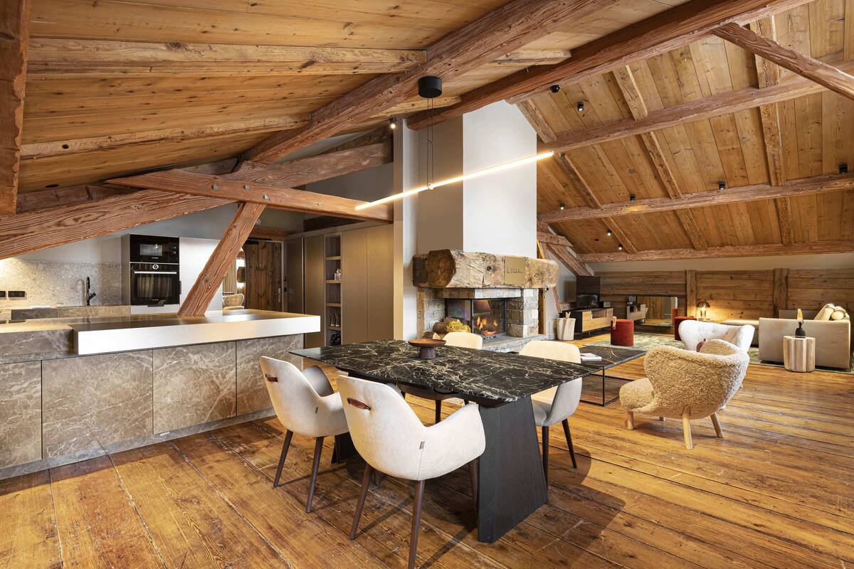 Um amplo loft com teto alto, vigas de madeira aparentes, área de jantar central com mesa de mármore preto, cadeiras brancas e lareira.