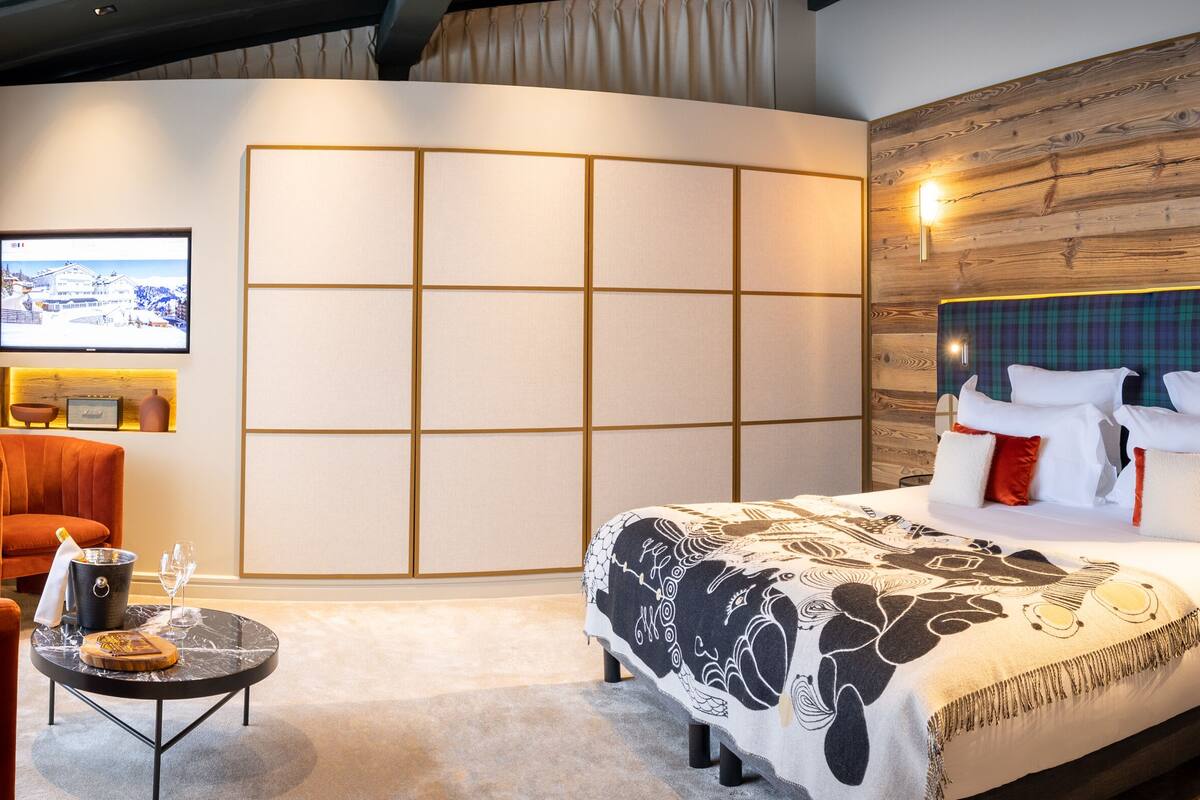 Quarto de hotel com uma cama grande, uma mesa pequena com uma garrafa e copos, uma cadeira e vista para uma paisagem nevada pela janela.