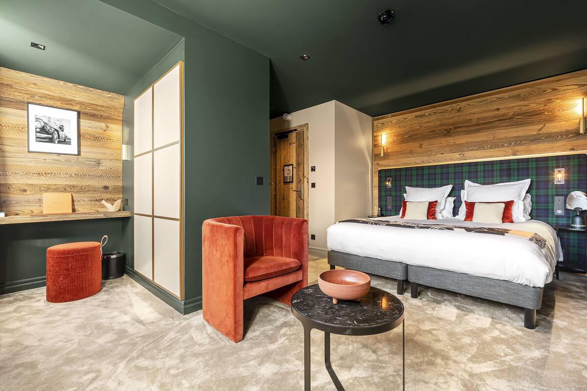 Quarto de hotel com cama, uma cadeira vermelha, uma mesinha e um painel de madeira decorativo com um quadro.