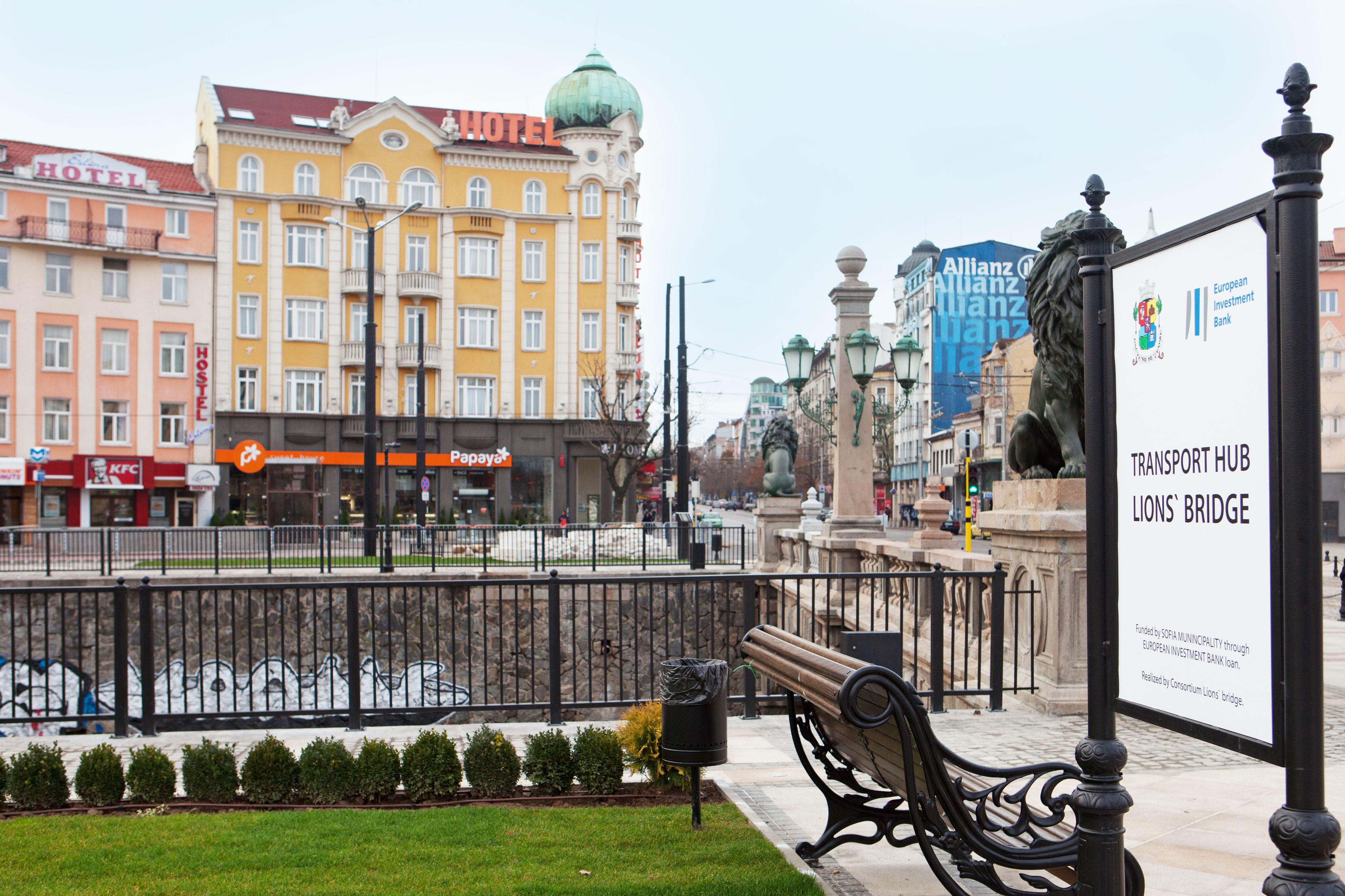 A city square with a bench, a statue, and a sign for the Transport Hub Lions' Bridge.