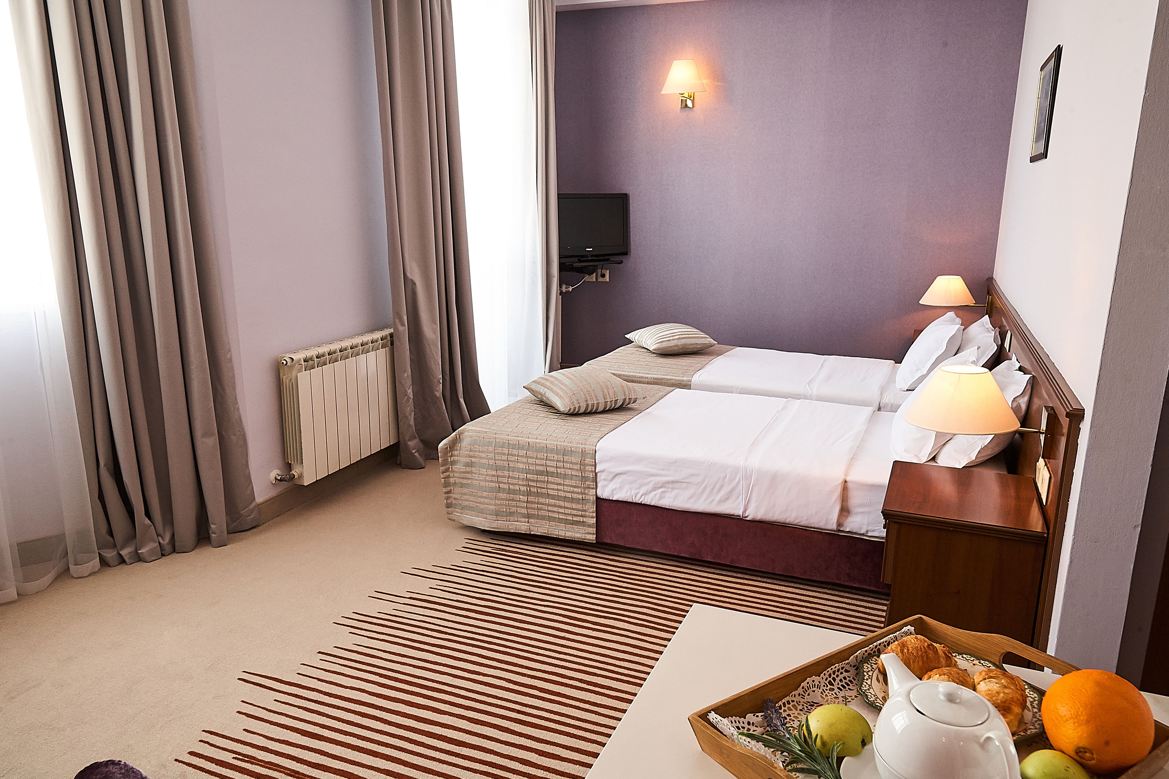 A hotel room with a bed, a bedside table with a lamp, a television, and a tray with fruits and a teapot.