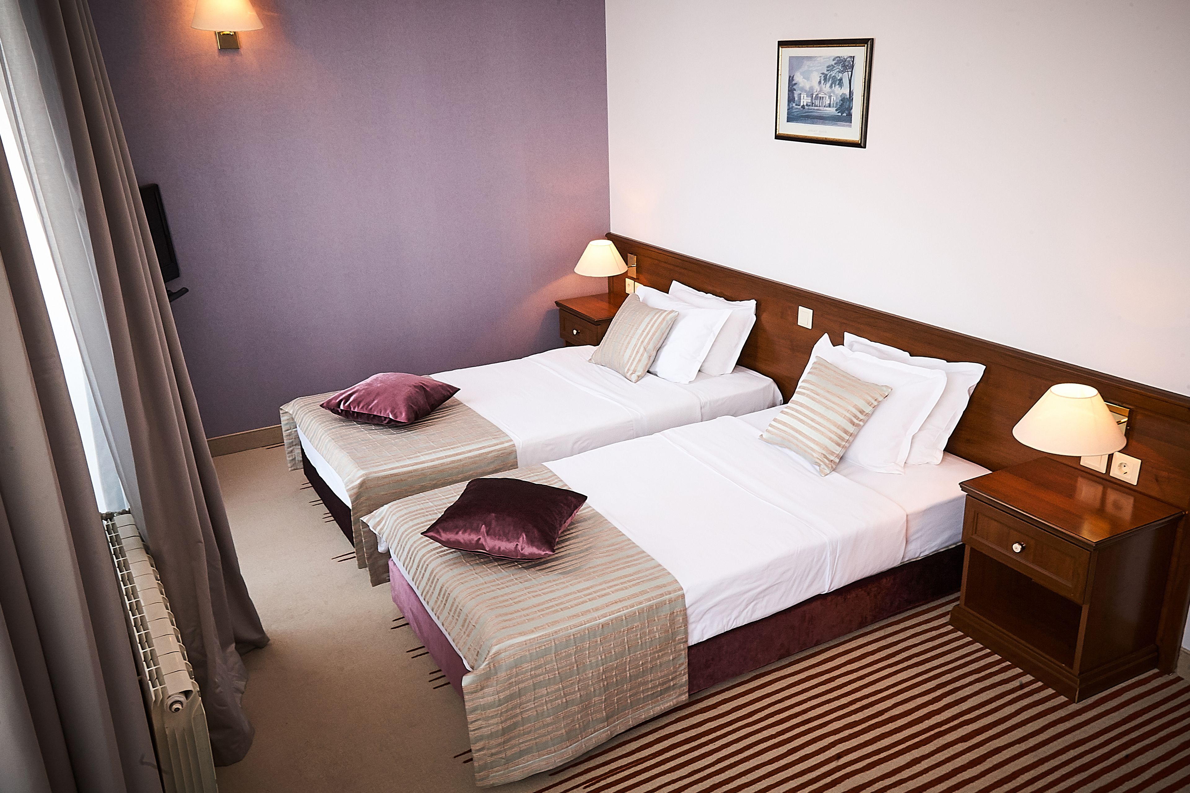 A hotel room with two beds, a nightstand with a lamp, a wall-mounted picture, and a radiator.
