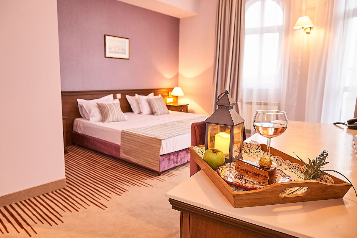 A hotel room with a neatly made bed, a bedside table with a tray of snacks, a wine glass, and a lantern.