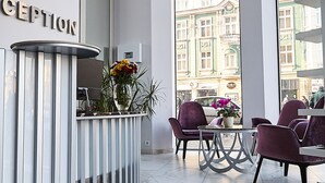 Uma área de recepção com balcão, cadeiras, um vaso com flores e uma janela grande com vista para edifícios.