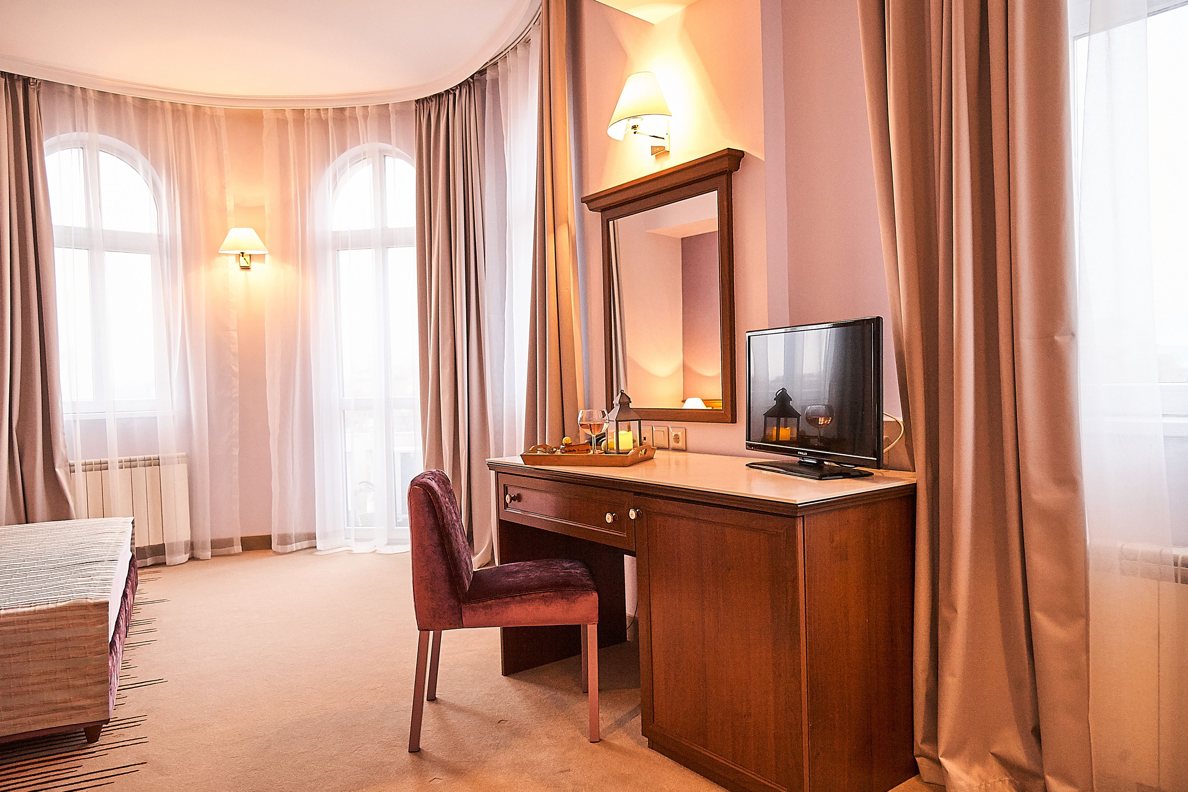 A hotel room with a wooden desk, a chair, a television, and two wall-mounted lamps.
