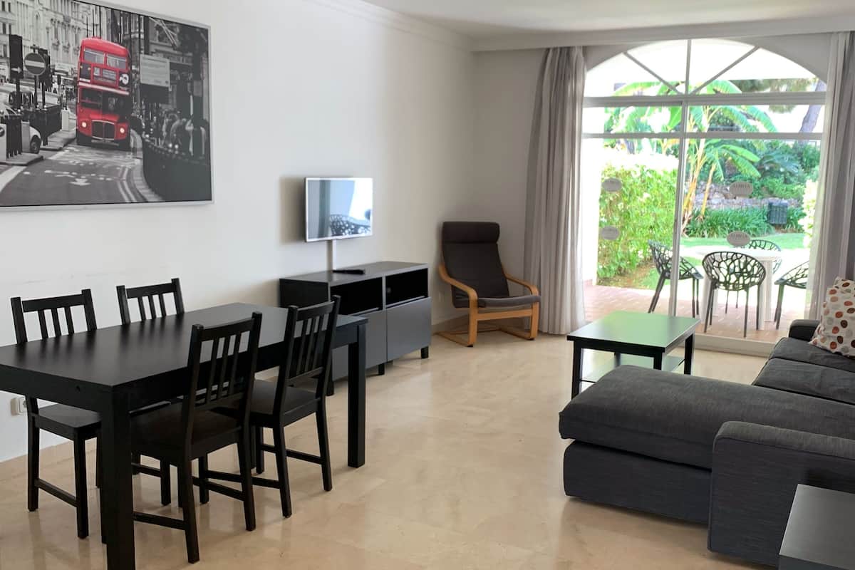 Een moderne woonkamer met een eethoek, een televisie en een groot raam met uitzicht op groen.