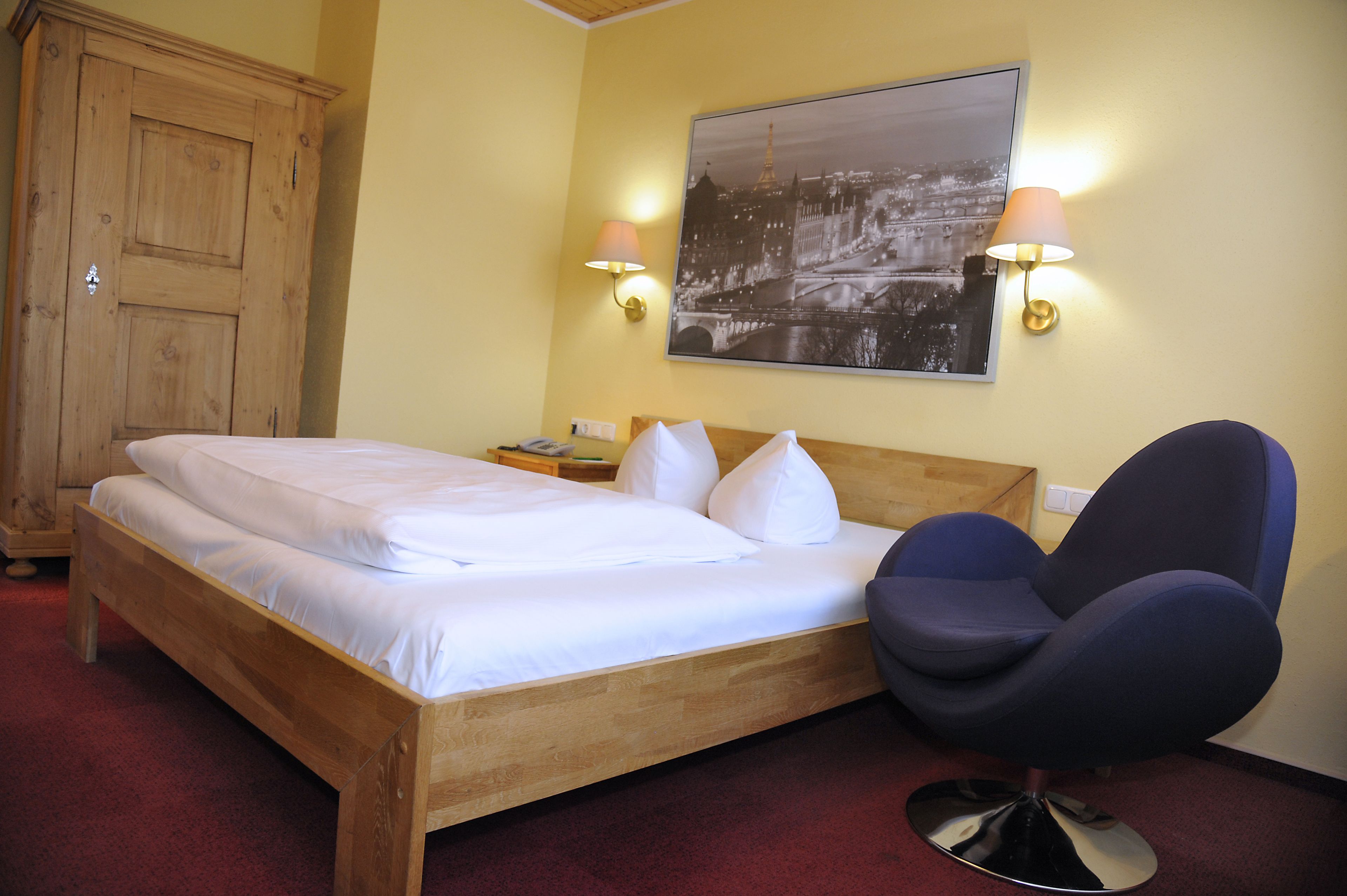 A hotel room with a wooden bed, two bedside tables, a chair, and a wall-mounted lamp.