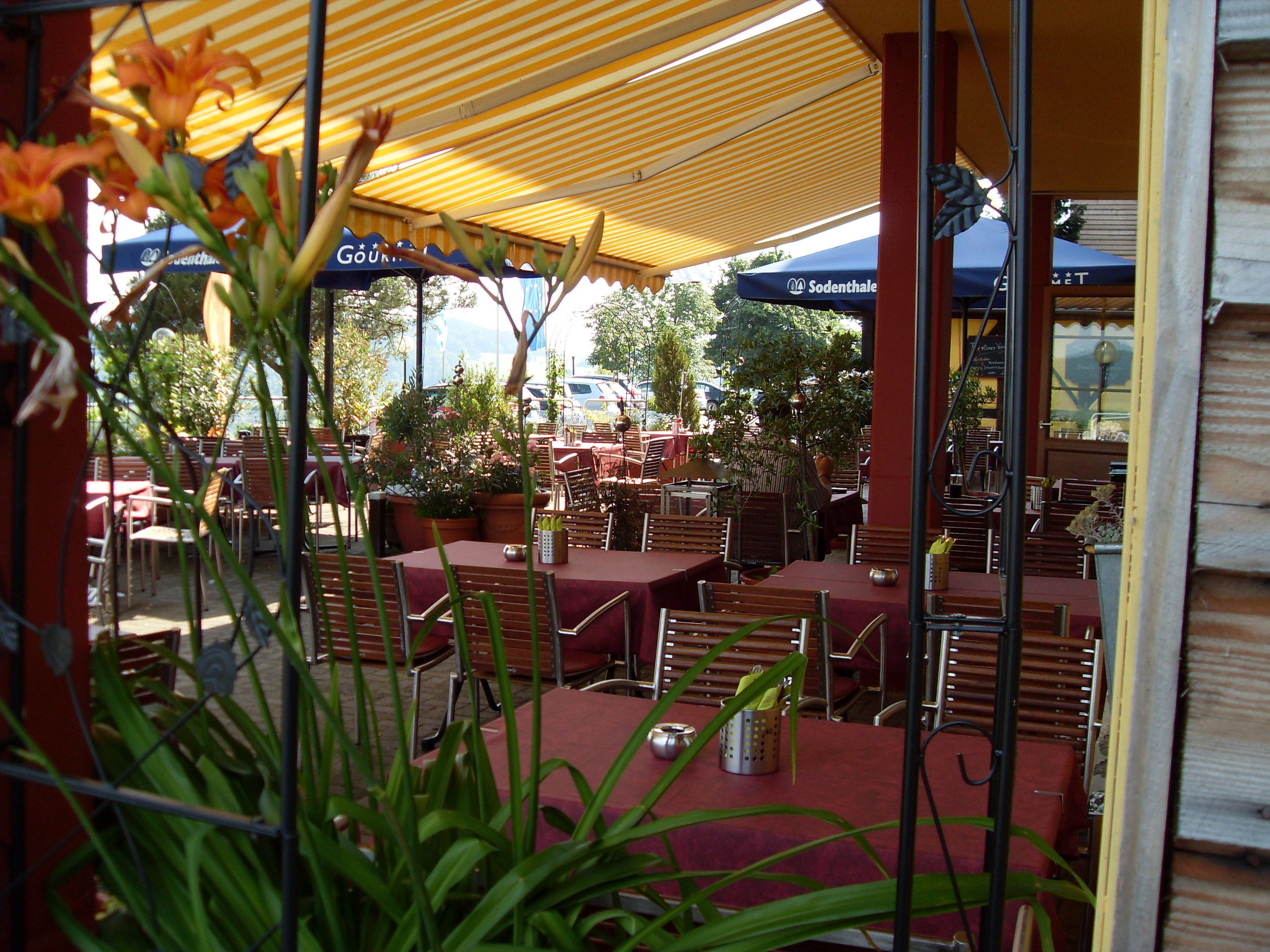 Un café al aire libre con mesas y sillas bajo toldos a rayas, rodeado de macetas con plantas.
