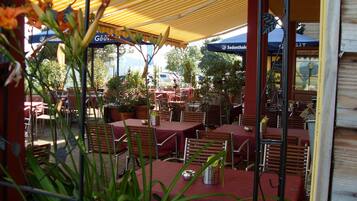 Un café al aire libre con mesas y sillas bajo toldos a rayas, rodeado de macetas con plantas.