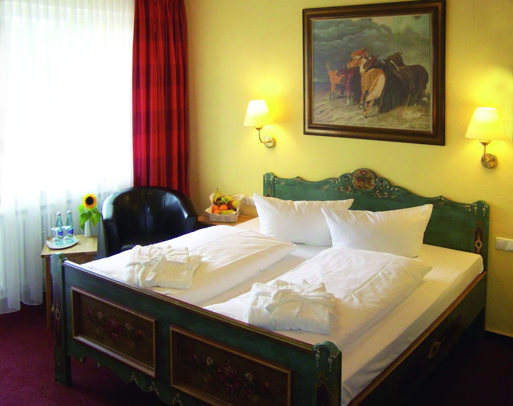A hotel room with a bed, two bedside lamps, a painting on the wall, a chair, a table with a fruit bowl, and a window with curtains.