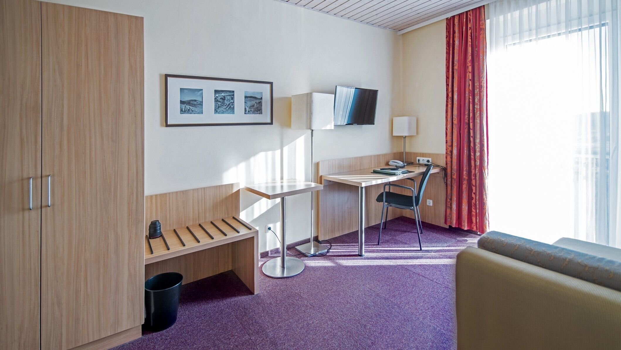 A hotel room with a desk, chair, and a bed. There is a window with curtains, a mounted TV, and a framed picture on the wall.