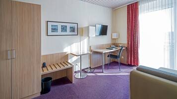 A hotel room with a desk, chair, and a bed. There is a window with curtains, a mounted TV, and a framed picture on the wall.