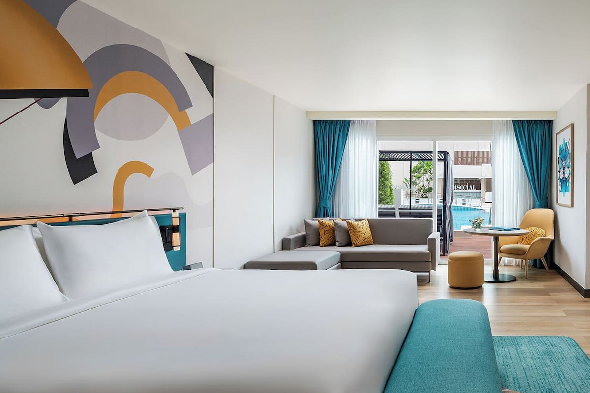 A modern hotel room with a large bed, a sofa, a small table, and a view of a pool.