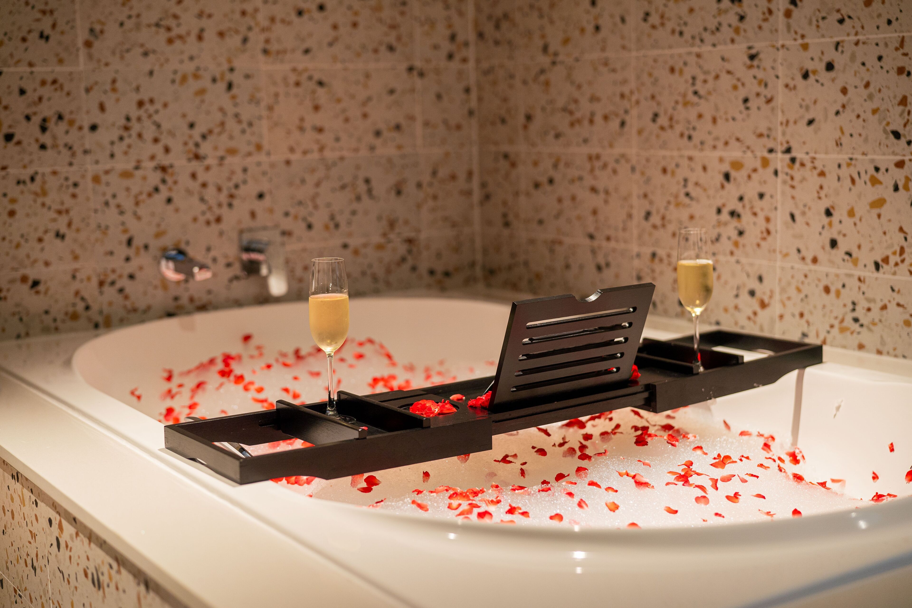 A bathtub with rose petals and two champagne glasses on a tray.