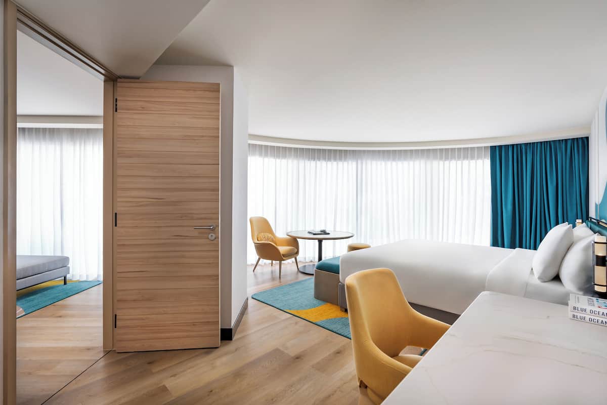 A hotel room with a bed, a desk, a chair, a sofa, a table, and a large window with curtains.