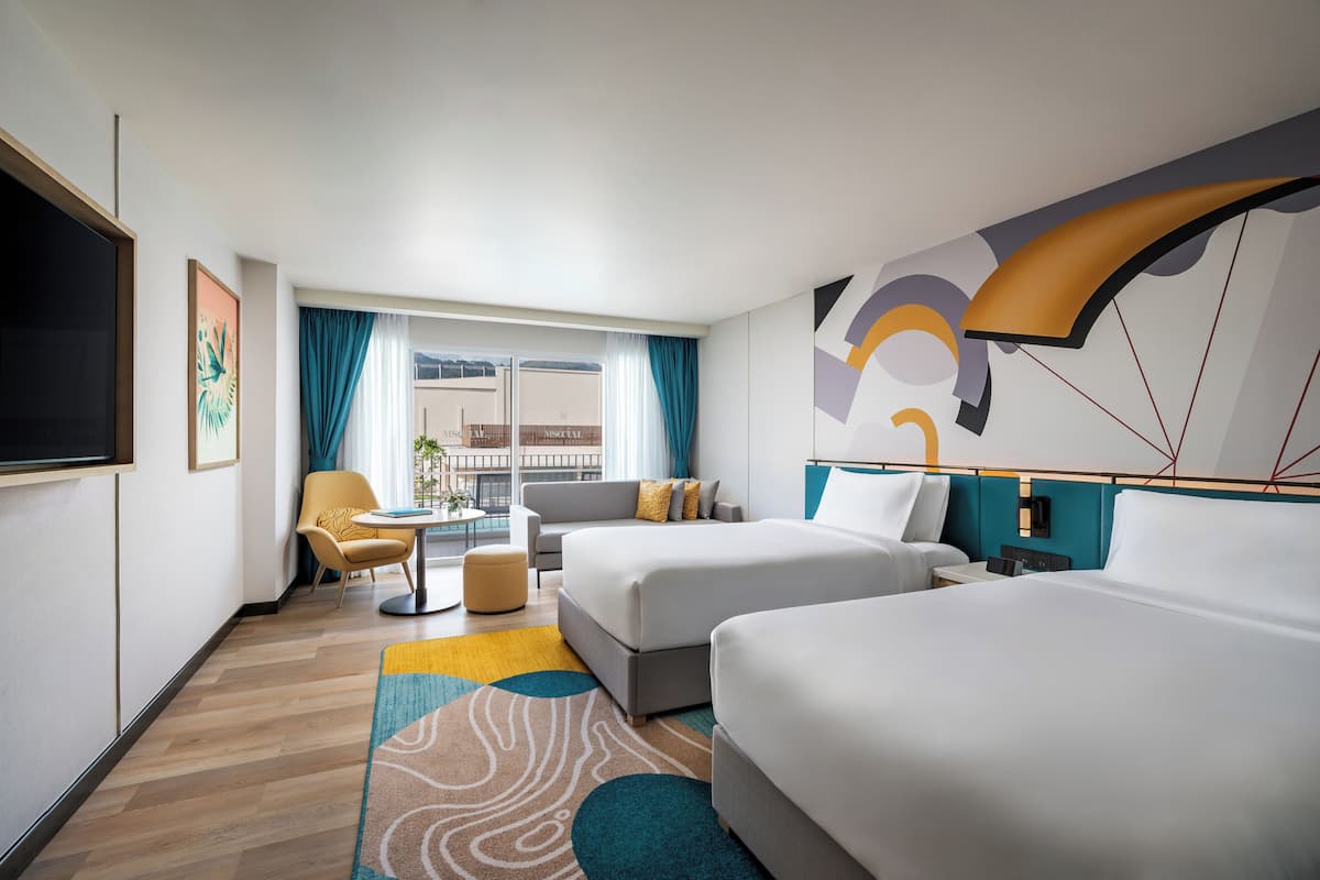 A hotel room with two beds, a sofa, a TV, a window with curtains, and a view of buildings outside.