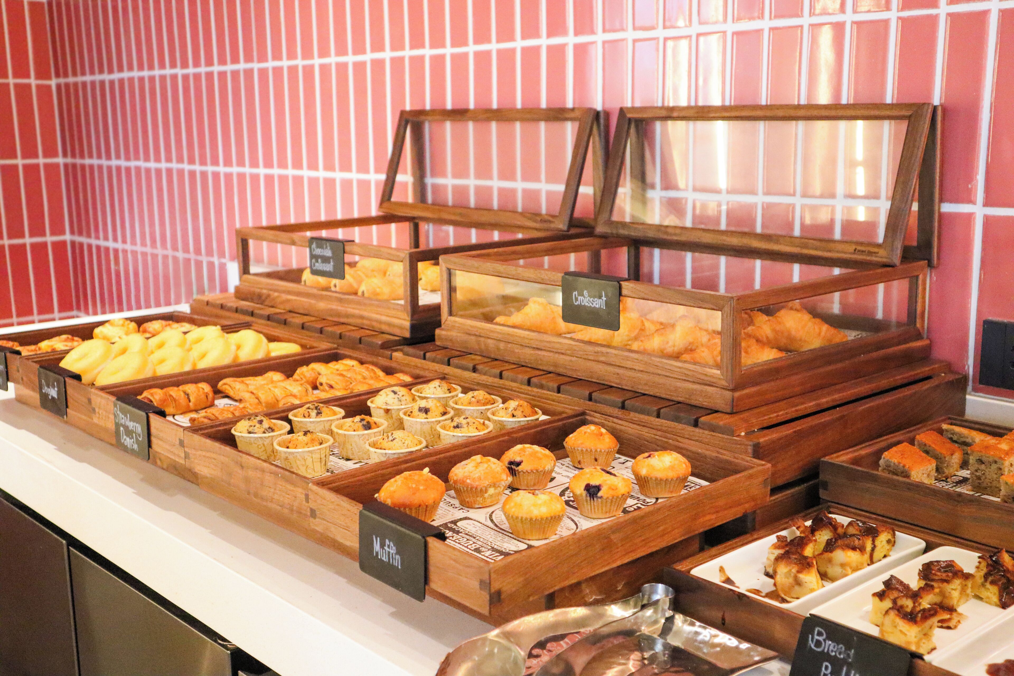 Eine Bäckereiauslage mit verschiedenen Backwaren in Holzkästen, beschriftet mit Namen wie ‚Croissant‘ und ‚Muffin‘.