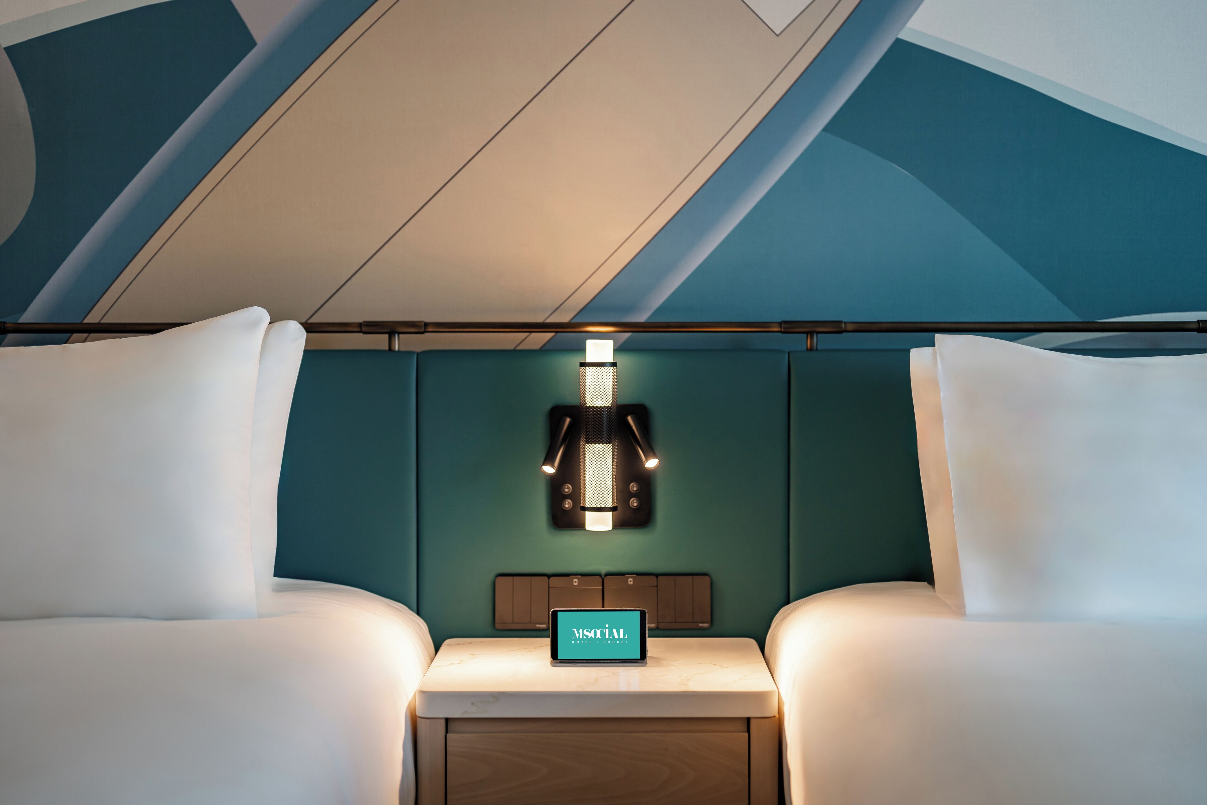 A hotel room with two beds, a nightstand with a digital clock, and a wall-mounted light fixture.