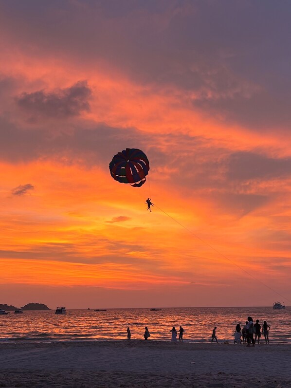A beach scene at sunset with a parasail in the sky, people on the shore, and boats in the water.